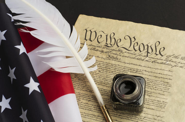 staged photo of copy of Constitution with a quill and bottle of ink and American flag beside of it