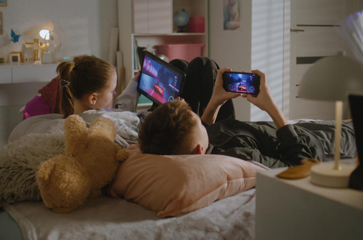 Three children lie on a bed watching a movie on a tablet, with a teddy bear nearby in a softly lit bedroom.