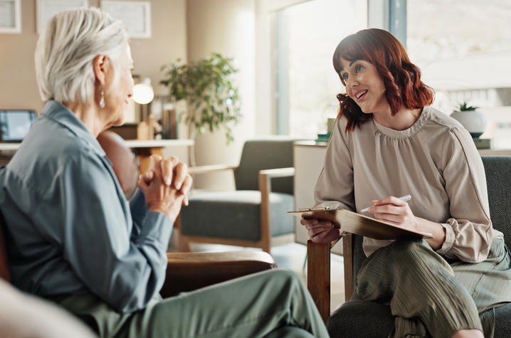Two people sit across from each other in a bright office, with one holding a clipboard and pen while talking with the other.