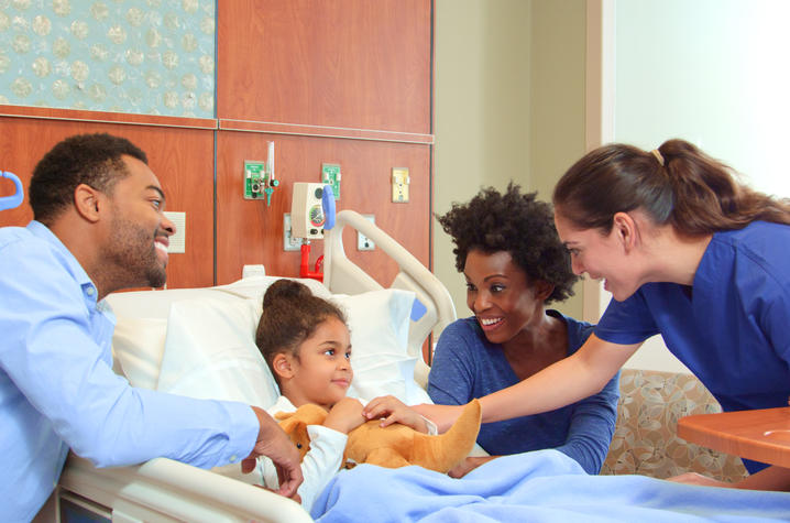 A child lies in a hospital bed while two adults sit nearby, and a health care provider stands at the bedside adjusting a blanket or comfort item. The room includes medical equipment and bedside furniture.