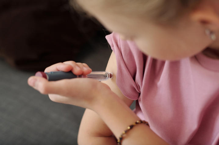 Close-up of a child in a pink shirt using an insulin pen to administer an injection into the upper arm.