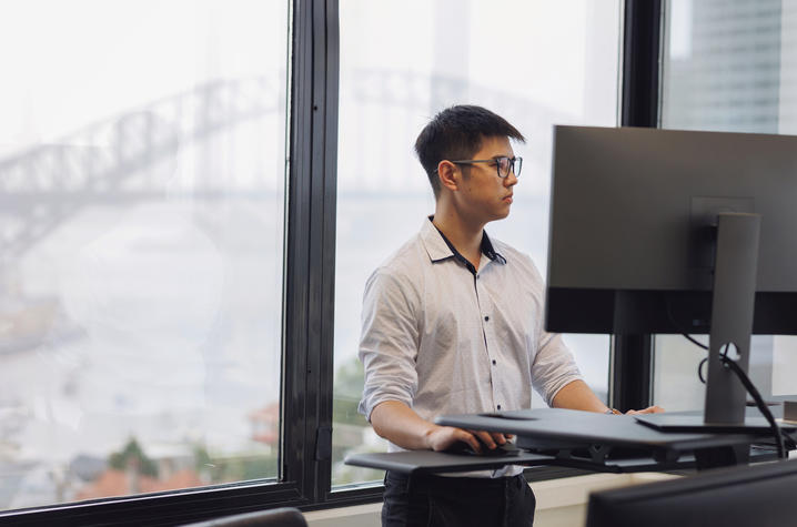 Person works at a standing desk in an office with floor-to-ceiling windows overlooking a harbor bridge and city skyline.