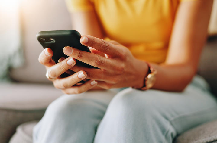 Person using a smartphone while seated on a couch. Close-up of a person seated on a couch holding and using a smartphone with both hands. The image focuses on the phone and hands, with a blurred indoor background.