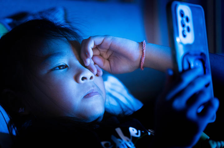 A child lies in bed in a dark room, holding a smartphone close to their face. Blue light from the phone illuminates the child's face and hand while the rest of the room remains dimly lit.