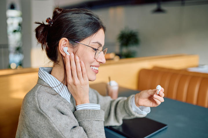 Person putting on wireless earbuds at a cafe table. Person wearing glasses sits at a table in a cafe, placing a white wireless earbud into one ear while holding the matching earbud in the other hand. A tablet rests on the table, with seating and soft lighting visible in the background.