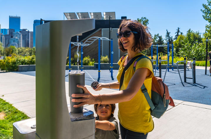 Stock photo showing someone refilling a water bottle at an outdoor fountain Person fills a reusable bottle at an outdoor water fountain on a sunny day while a child stands nearby in a park setting.