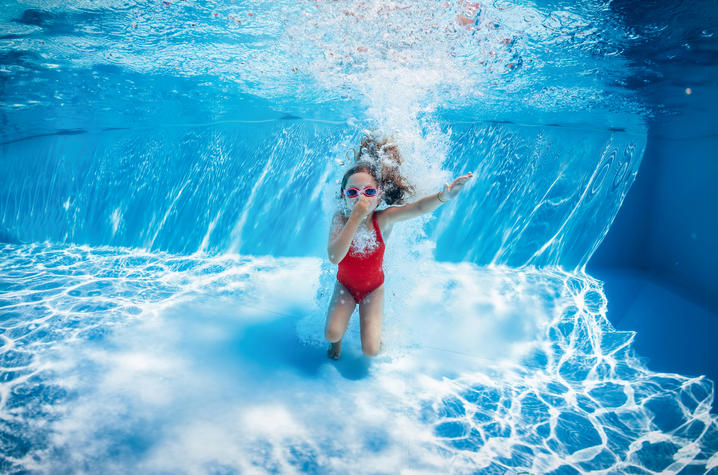 Stock photo of child swimming underwater in pool Child swimming underwater in a pool, wearing goggles and a red swimsuit, surrounded by bubbles and blue water.