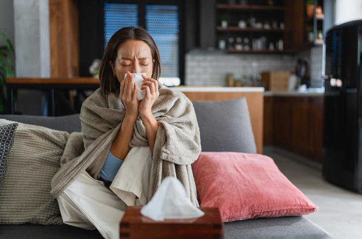 Person with cold symptoms resting at home with tissues. Person wrapped in a blanket sits on a couch at home holding a tissue to their nose. A tissue box rests on a table in the foreground, suggesting cold or respiratory illness symptoms.
