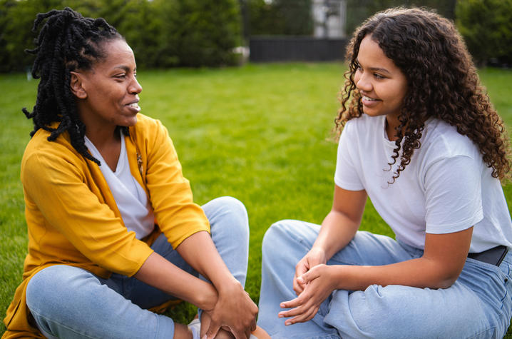 Stock photo of mother with adolescent child A parent and teen daughter sit cross-legged on a grassy lawn, smiling and talking face-to-face during an outdoor conversation.