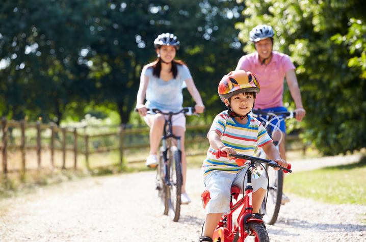 A child wearing a helmet rides a red bicycle along a sunlit gravel path, with two helmeted adults riding bikes behind them in a wooded park setting.
