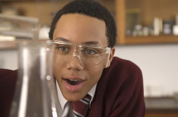 A student wearing safety goggles leans in closely, observing a beaker during a science experiment. Their focused expression suggests curiosity and engagement in a classroom or lab setting.