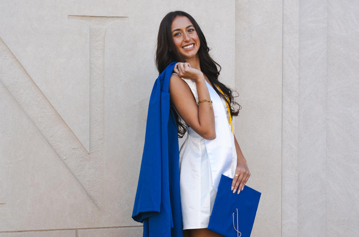 Graduate in a white dress holds a blue cap with a gown over one shoulder against a stone wall.
