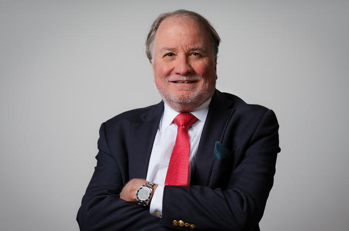 Photo of Greg Gerhardt Studio portrait of a mature adult in a dark suit with a white shirt and bright red tie, arms crossed. A silver watch is visible on the wrist and a patterned pocket square in the jacket. Neutral light gray background with even lighting.