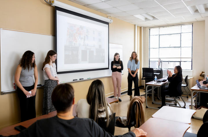 Four women stand at the front of a classroom beside a projected slide, appearing to present to seated students; desks, notebooks, and a computer workstation are visible, with large windows letting in daylight.
