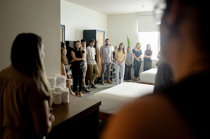 A group of adults stands closely together in a modern hotel room or suite, facing inward as if listening to someone speaking off-camera; a bed and countertop are visible, and the scene is viewed from behind two blurred figures in the foreground.