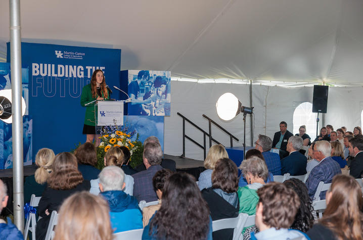 Speaker stands at a podium addressing a seated audience under a tent, with blue “Building Together” signage and presentation slides visible in the background.