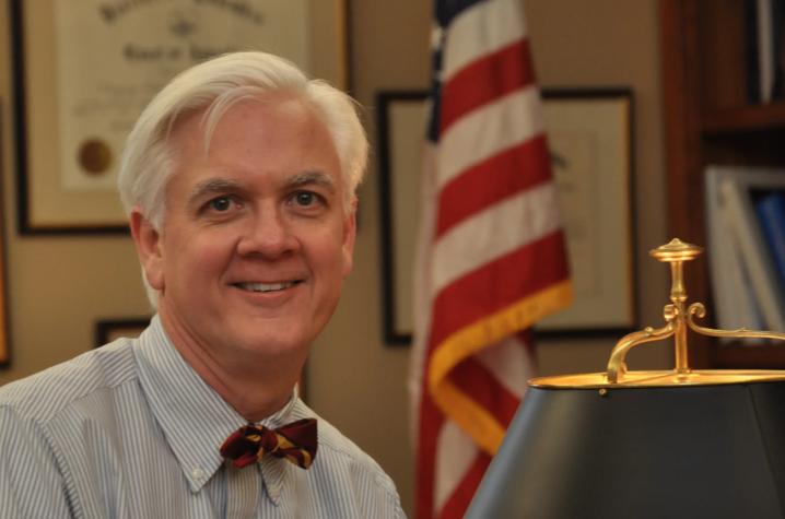 Person seated at a desk in an office, wearing a dress shirt and bow tie, with an American flag and desk lamp visible in the background.