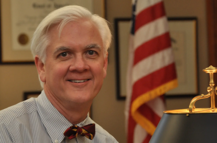 Person seated at a desk in an office, wearing a dress shirt and bow tie, with an American flag and desk lamp visible in the background.