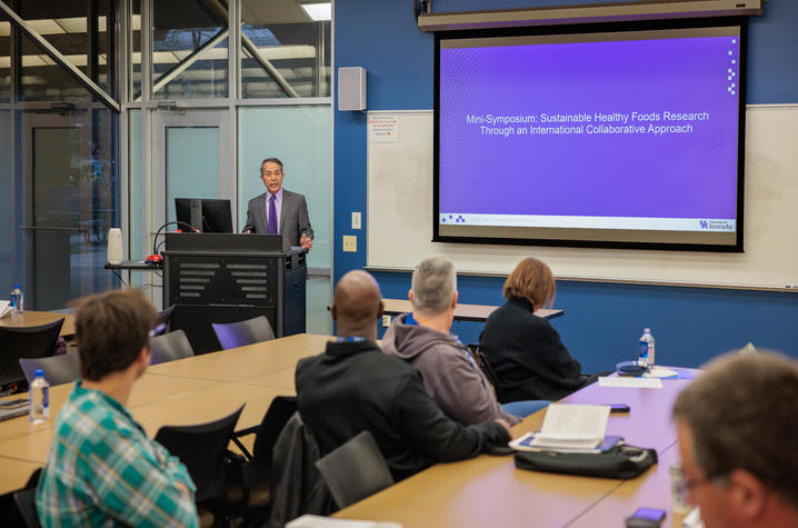 A presenter stands at a podium at the front of a classroom-style lecture space. The presenter is positioned slightly to the left, behind a black lectern with a computer monitor on top. Several people sit at tables facing the front of the room. 