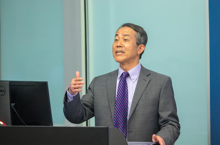 Person stands at a podium, appearing to give a presentation. The person is wearing a patterned suit jacket, a collared shirt, and a striped tie. One hand is raised in a gesturing motion while the other rests near the podium.