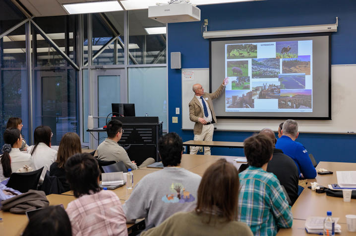 A group of people sits at tables in a classroom-style room, facing a presenter at the front. The presenter stands near a projection screen and points to a slide displaying multiple photographs. The presenter stands beside a podium with a computer monitor.