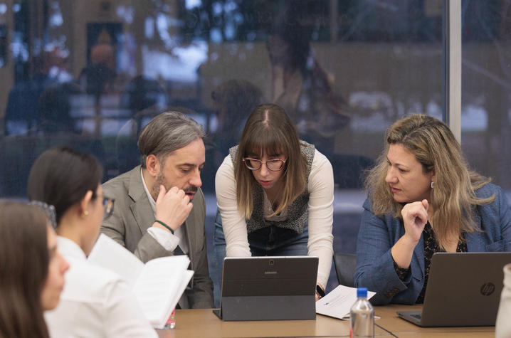 A small group of people sits and stands around a table, looking at two open laptop computers. One person seated on the left holds a notebook with pages open. Another person in the center leans forward toward one of the laptops, while two people sit.
