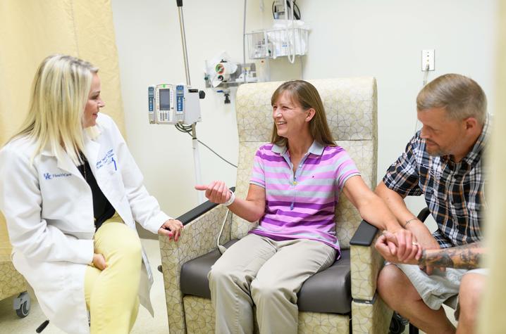 A young nurse in a white coat sits beside a middle-aged white woman in a hospital chair; her husband is sitting beside her and holding her hand. 
