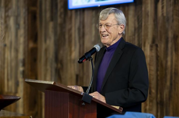 A person stands at a wooden podium speaking into a microphone, wearing a dark blazer and purple shirt, with a rustic wooden wall and a screen in the background.