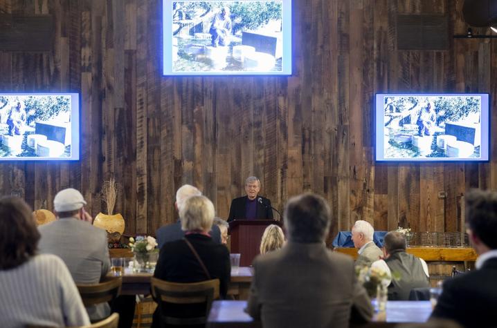 Audience seated at tables listening to a speaker at a podium in a rustic venue with three large screens displaying the same image on a wooden wall.