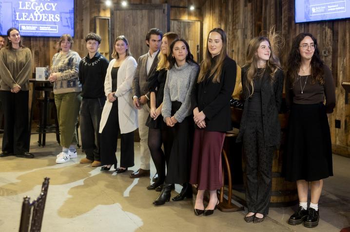 A group of people stands in a line inside a rustic venue with wooden walls. Three large screens are visible in the background, displaying text and images. A podium is partially visible on the right.