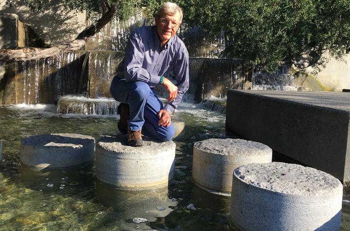 Horst Schach posing for A person kneels on a large round stone in a shallow water feature with other circular stepping stones, surrounded by greenery and a small waterfall in the background.a picture at the Levi's Plaza in San Francisco