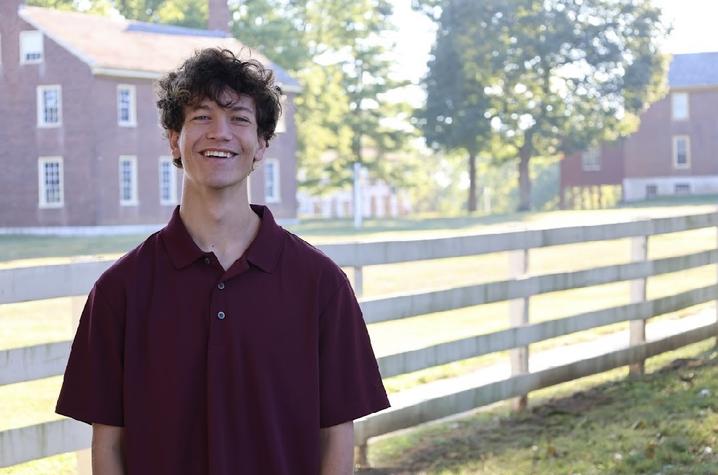 Person standing outdoors in front of a wooden fence and brick buildings, wearing a maroon short-sleeve polo shirt, with trees and grass in the background.