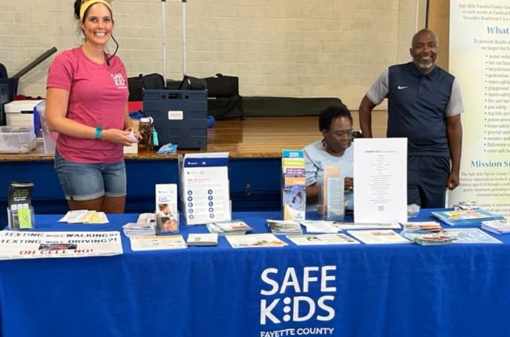 image of three people at table with blue tablecloth that says "Safe Kids Fayette County"