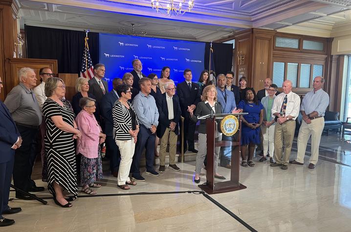 image of people standing behind Ellen Hahn as she speaks from a podium