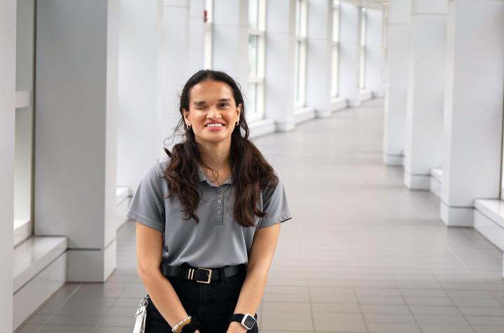 A person stands in a bright, modern hallway with large windows, smiling at the camera. They are wearing a gray polo shirt, dark pants, and a smartwatch, with keys clipped at their side.