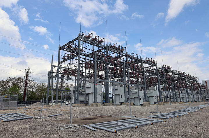 Photo of electrical substation Wide-angle view of a large electrical substation with steel support structures, transformers, and high-voltage equipment extending across a gravel-covered site beneath a blue, partly cloudy sky.