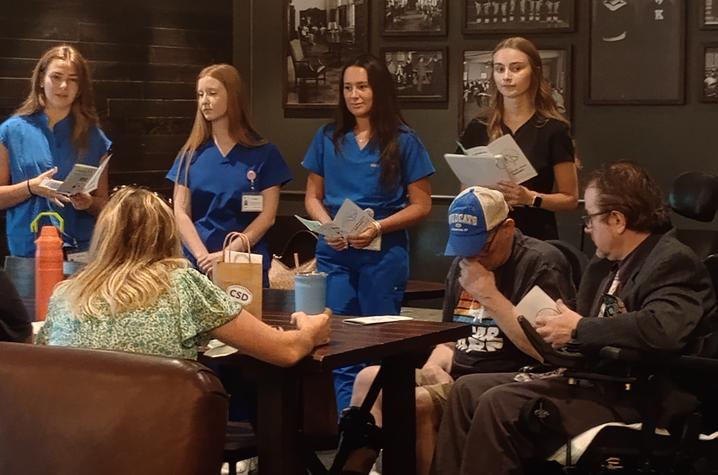 Photo of Aphasia Lab students at Starbucks Participants seated around a conference table listen as four individuals in blue scrubs stand at the front of a meeting room holding papers and presenting. Framed photos hang on the wall behind them.