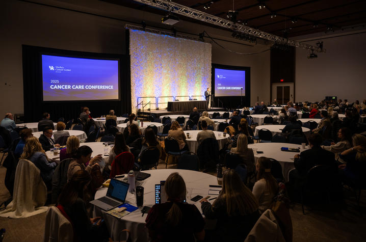 Attendees at round tables facing a stage where speakers present at the 2025 Cancer Care Conference hosted by UK Markey Cancer Center, with illuminated backdrop and multiple display screens.