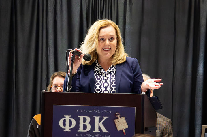 Person speaking at a podium with a microphone, standing in front of a Phi Beta Kappa banner and black curtain.