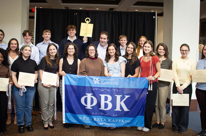 Group of people standing indoors holding certificates behind a blue Phi Beta Kappa banner.