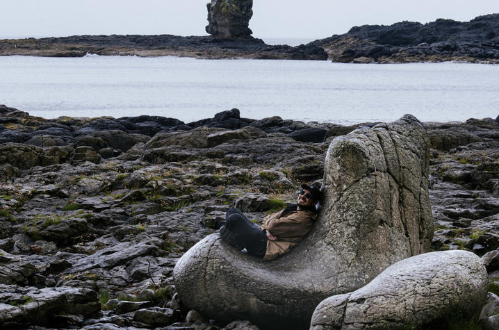 photo of Christian Dordelly A person sits on a large rock formation along a rugged shoreline during a visit to a coastal landscape.