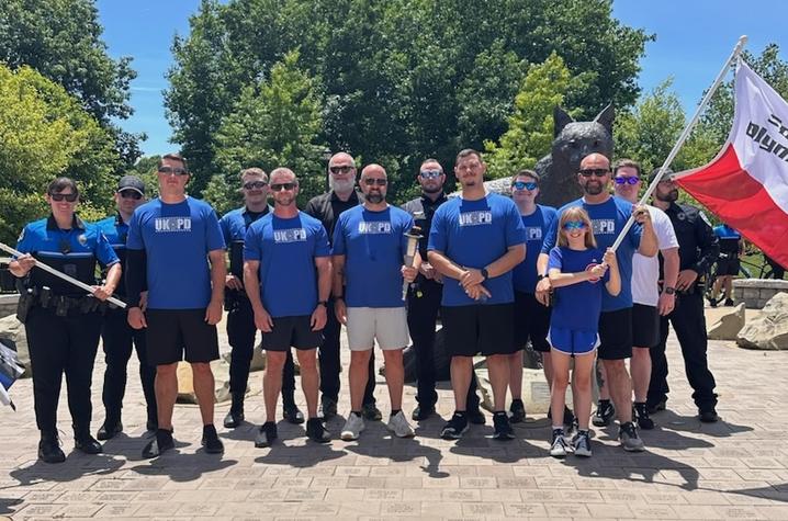 A photo of the UKPD torch run volunteers from 2024 standing in front of the Bowman statue on campus. Capt. Adams is in the center of the group, they're all wearing blue UKPD shirts. They’re holding a red and white Special Olympics flag and a UKPD flag.