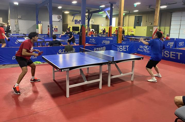 Two people play an intense game of table tennis in a large indoor facility with multiple courts and blue barriers.