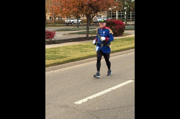 Brian Rice runs A man dressed in a blue Prince Charming-style costume runs down a road during a marathon, with autumn trees and landscaped bushes in the background.