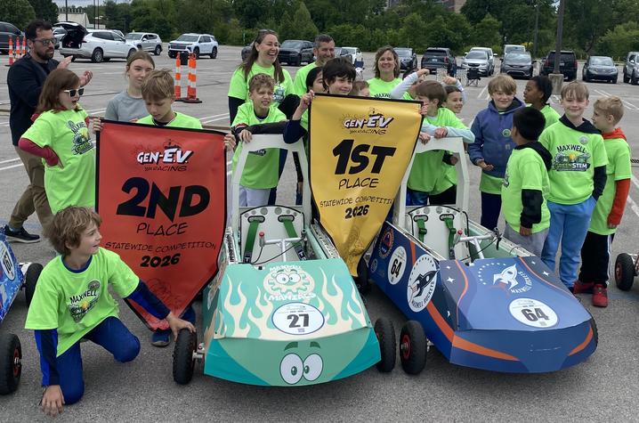 Maxwell Spanish Immersion Elementary teams. Students wearing green shirts celebrate around their race cars while holding banners for first- and second-place finishes at the GEN‑EV statewide competition.