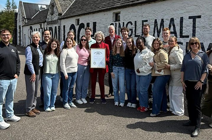 A group of about 20 people stand outdoors in front of a white stone building with large black painted lettering on the wall. One person in the center holds a framed certificate while the group gathers around for a photo.