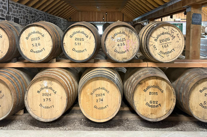Rows of large wooden whisky barrels are stacked on wooden racks inside a distillery aging warehouse with exposed timber beams and stone walls. The barrel ends are labeled “Ballindalloch Distillery.”