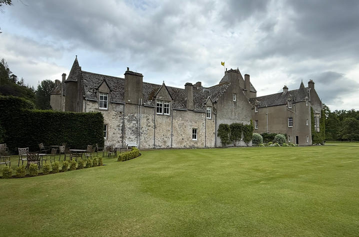 Large historic stone manor house with steep slate roofs, chimneys, and turreted architectural details, viewed from a wide, manicured lawn. Outdoor tables and chairs sit on a landscaped patio area to the left, bordered by tall hedges and trees.