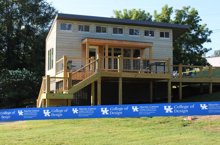 A wooden modular emergency housing prototype with a raised deck and stairs sits on a grassy property.