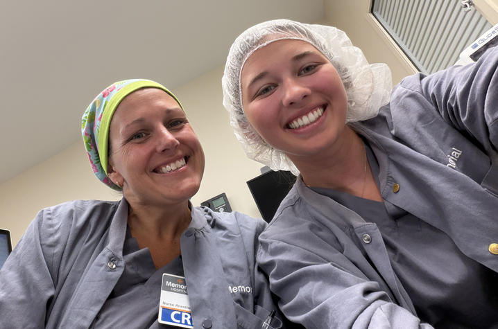 Maria Morris shadowing a CRNA Two people in gray medical scrubs and protective hair coverings pose for a close-up photo in a clinical setting.